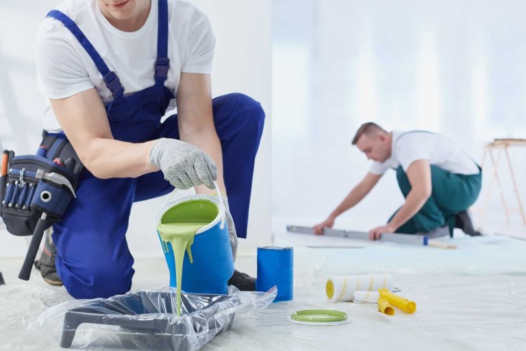 Due operai in camice bianco e tuta sono impegnati con ristrutturazioni complete, dipingendo una stanza. Uno versa vernice verde chiaro in una vaschetta mentre l'altro misura sul pavimento. Rulli e secchi li circondano, con teli di plastica che coprono ordinatamente il pavimento.