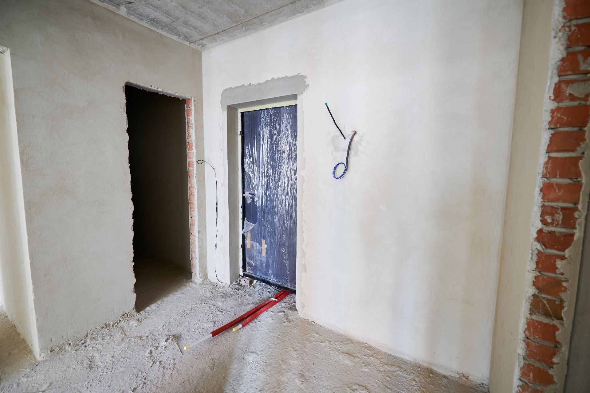 L'interno di un edificio in fase di ristrutturazioni complete presenta pareti grezze con mattoni a vista e superfici parzialmente intonacate. Un portone, coperto di plastica, e materiali da costruzione sparsi evidenziano la trasformazione in corso.