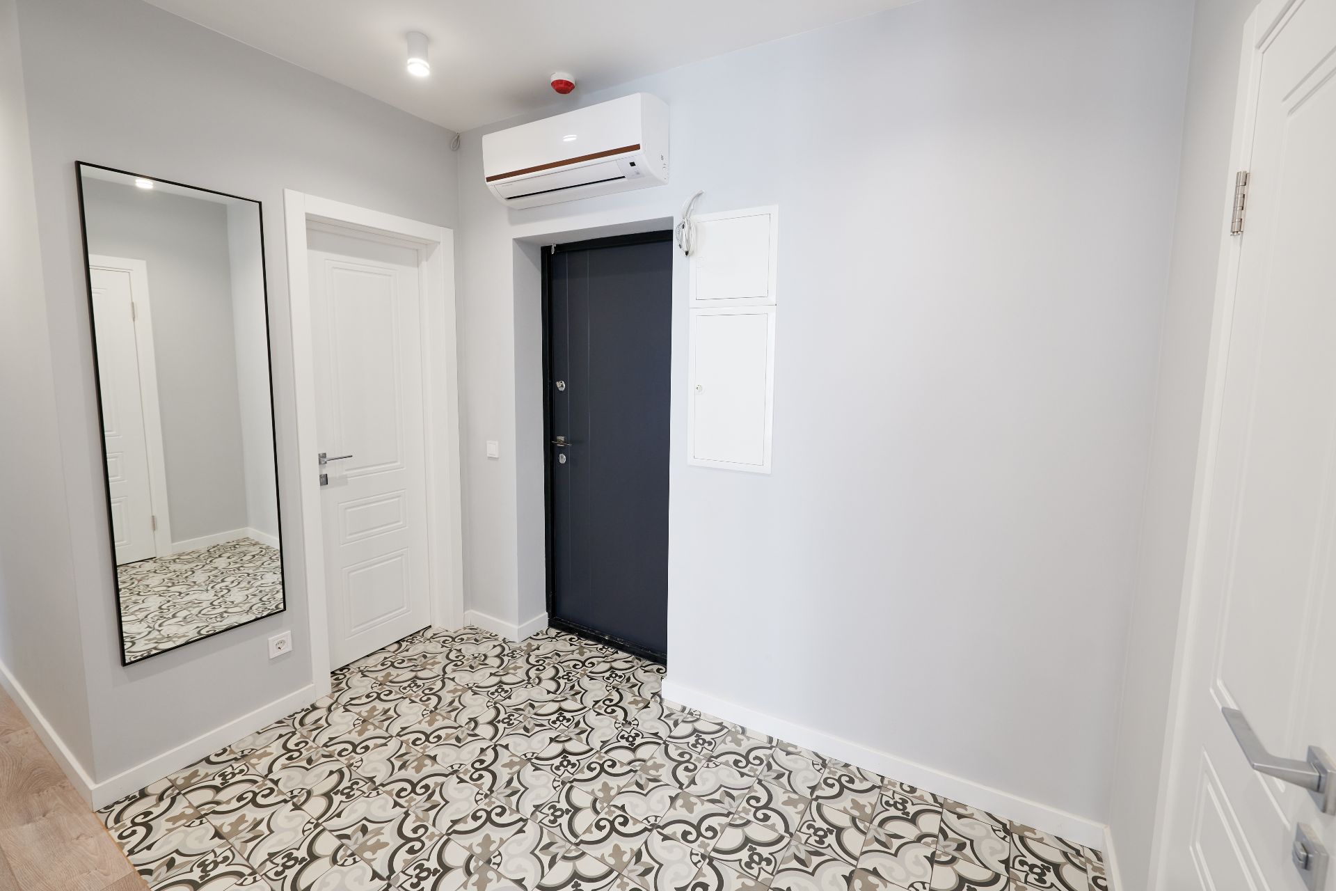 La stanza bianca, parte di ristrutturazioni complete, vanta un pavimento in piastrelle a motivi e un grande specchio sulla sinistra. Presenta una porta bianca chiusa, una porta nera con un condizionatore sopra, pareti grigio chiaro ed è illuminata da una plafoniera.