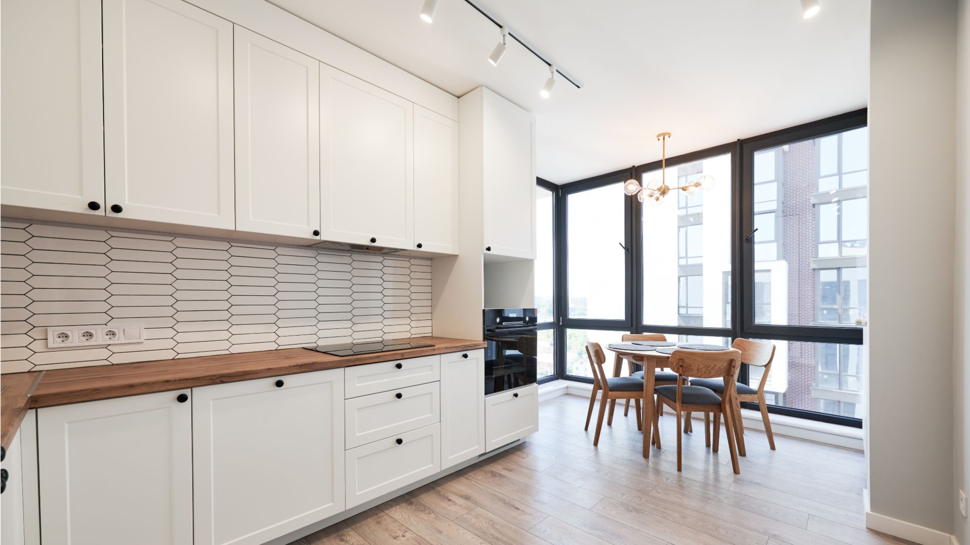 Cucina moderna con mobili bianchi, ripiani in legno e paraschizzi in piastrelle esagonali. La zona pranzo, con tavolo e sedie in legno, è vicina a grandi finestre. Questo progetto ristrutturazioni chiavi in mano è completato da un'elegante lampada da soffitto per uno spazio luminoso e contemporaneo.