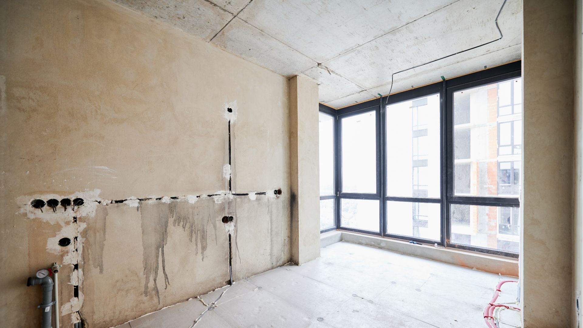 Una stanza incompiuta con pareti di cemento nudo, tubature esposte e prese elettriche attende la sua trasformazione. Le grandi finestre sulla destra lasciano entrare la luce naturale, rivelando una vista sulla città. Questo spazio è maturo per "ristrutturazioni chiavi in mano" per sbloccare il suo pieno potenziale.
