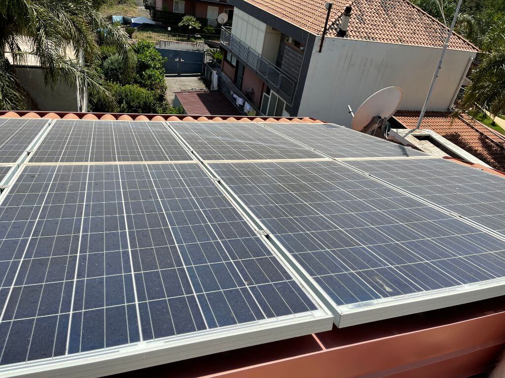 Una vista sul tetto mostra diversi pannelli solari installati su un tetto di tegole rosse in mezzo a lavori edili in corso, con uno sfondo di alberi ed edifici residenziali. I pannelli sono inclinati in modo ottimale per catturare la luce solare, contribuendo a una vita sostenibile.